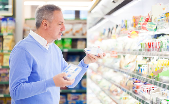 Herr mit grauen Haaren, weißem Hemd und hellblauem Pullover steht vor einem Kühlregal in einem Supermarkt