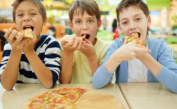 Drei junge Buben haben jeweils eine Pizzaschnitte in der Hand und beißen gerade genussvoll ab