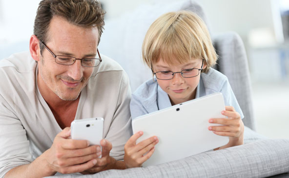 Papà e figlio con gli occhiali, sdraiati sul divano che consultano cellulare e tablet