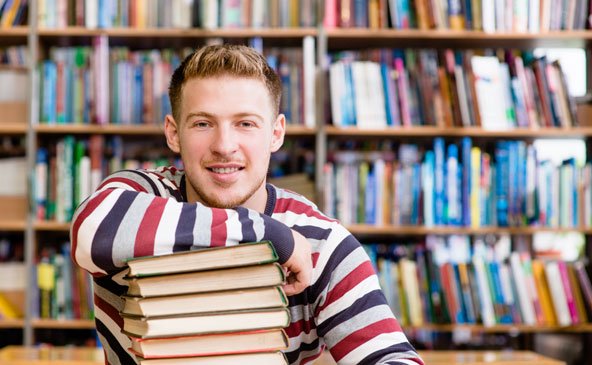 Junger Mann in einer Bibliothek lahnt einen Armen lässig auf einem Stapel von Büchern